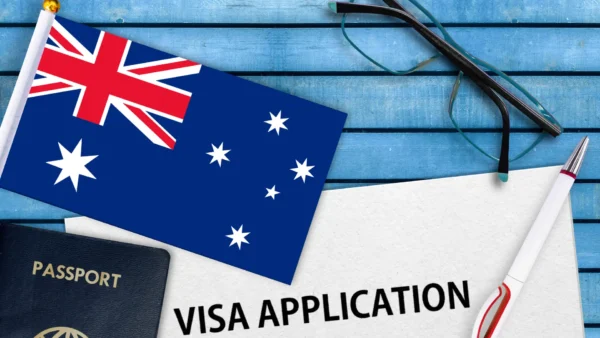 An Australian flag, a pair of glasses, a pen, a passport, and a document labeled Visa Application are arranged on a blue wooden surface.