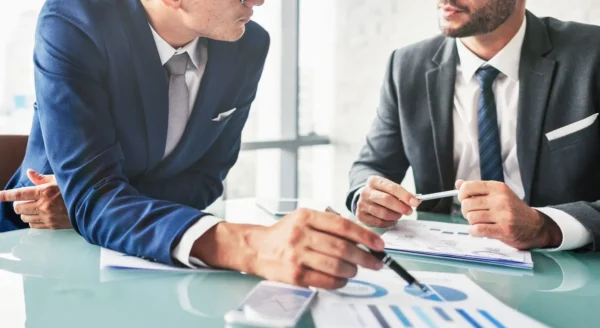 Two business professionals in suits reviewing printed charts and graphs during a meeting at a glass conference table. One person points to a pie chart with a pen while the other holds a pen and listens.