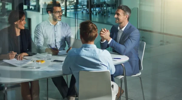 Four people sit around a table in a modern office, engaged in a meeting, with documents and glasses of water in front of them.