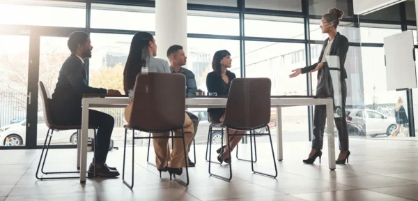 A woman stands and speaks to four seated colleagues in a modern office meeting room with large windows.