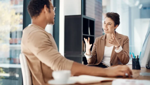 A woman in a blazer gestures animatedly while speaking to a man seated across from her at a desk, suggesting an engaged conversation or meeting in a bright office setting.