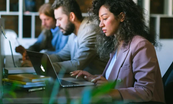 A woman with curly hair, wearing a blazer, sits at a desk focused on her laptop, with a stack of books beside her. Two other people are seated behind her, also working on laptops in a shared office or study space.