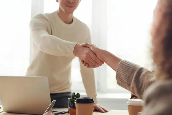 Two people are shaking hands across a desk with a laptop, coffee cups, and a small potted plant, suggesting a positive meeting or agreement in a bright, modern office setting.