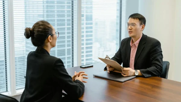 Two people in business attire sit across a table in an office, engaged in a conversation. One holds a clipboard and appears to be interviewing the other.